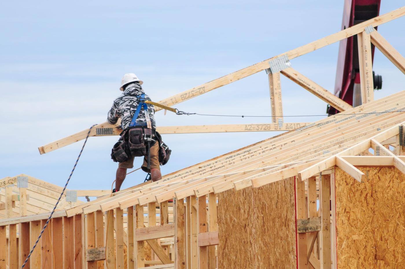 A construction worker installs wooden trusses on the roof of a building under a blue sky.