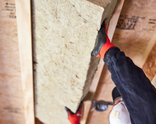 Worker installs fire-resistant fiberglass insulation between wooden wall studs during construction.