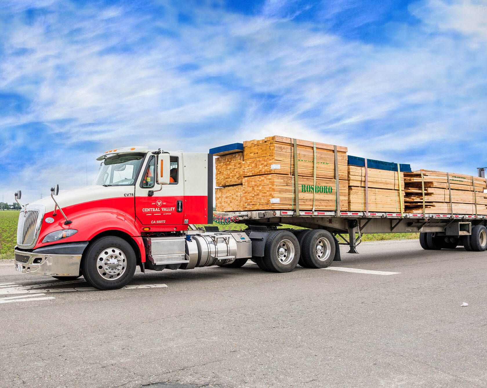 A red and white delivery truck labeled "Central Valley" hauling stacked lumber marked "ROSBORO" on a paved road under a blue sky.