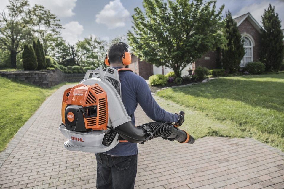 Man uses orange and gray Stihl BR 800 leaf blower on brick path in front of house.