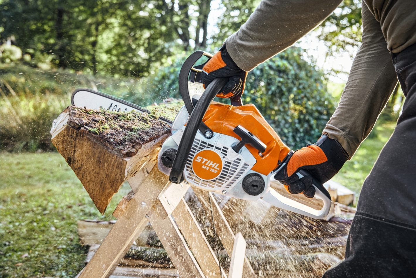 A person wearing gloves operates a Stihl MS 182 chainsaw to cut a mossy log on an outdoor sawhorse, with wood chips flying in the air.